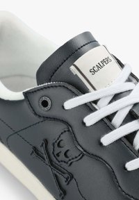 Schwarze Ledersneaker mit weißen Schnürsenkeln, strukturiertem Material und eingeprägtem Totenkopf- und Knochen-Design. Das Etikett trägt die Aufschrift "SCALPERS."