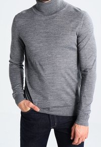 Maglione grigio a collo alto realizzato in morbido tessuto a maglia, con maniche lunghe, polsini a costine e una vestibilità aderente. Indossato con jeans scuri.