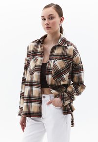 Koszula flanelowa w beżowo-brązowo-białą kratę z kołnierzykiem, dwoma przednimi kieszeniami i długimi rękawami, zestawiona z białymi spodniami.