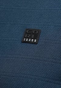 Σκούρο μπλε υφασμάτινη υφή με μαύρη τετράγωνη ετικέτα που γράφει "CORE JACK & JONES" με ανάγλυφα γράμματα.