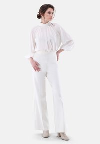 Blusa bianca con collo alto e dettagli plissettati, abbinata a pantaloni bianchi a gamba larga con vita alta e stivali con tacco beige. Tessuto morbido.