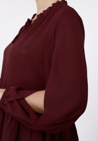 Μπορντό μπλούζα με βολάν στο ντεκολτέ, υφή υφάσματος και φαρδιά μανίκια με κοψίματα στους αγκώνες.
