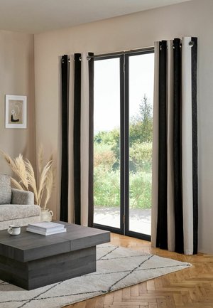 Wohnzimmer mit hohen Glastüren, schwarzen und beigen Vorhängen, grauem Sofa, dunklem Couchtisch, hellem Teppich und einer Vase mit Pampasgras an der Wand.