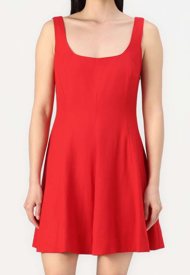 Femme portant une robe rouge vif sans manches avec un décolleté carré et une jupe évasée sur un fond uni.