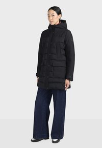 Femme portant une doudoune noire à capuche, un jean large bleu foncé et des chaussures noires, debout sur un fond clair uni.