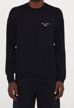 Mann trägt schwarzen langärmeligen Emporio Armani Milano Pullover mit kleinem weißem Logo auf der linken Brust, kombiniert mit schwarzen Hosen.