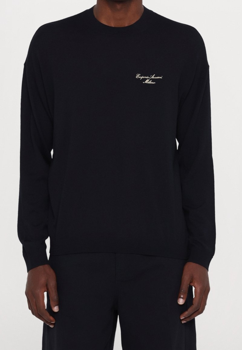 Homme portant un sweat à manches longues noir Emporio Armani Milano avec un petit logo blanc sur la poitrine gauche, assorti à un pantalon noir.