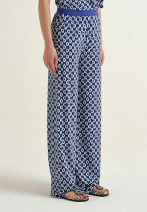 Donna che indossa pantaloni a zampa larga a motivo a diamante blu e bianco con cintura blu, abbinati a sandali con anello per il dito decorati con perline.