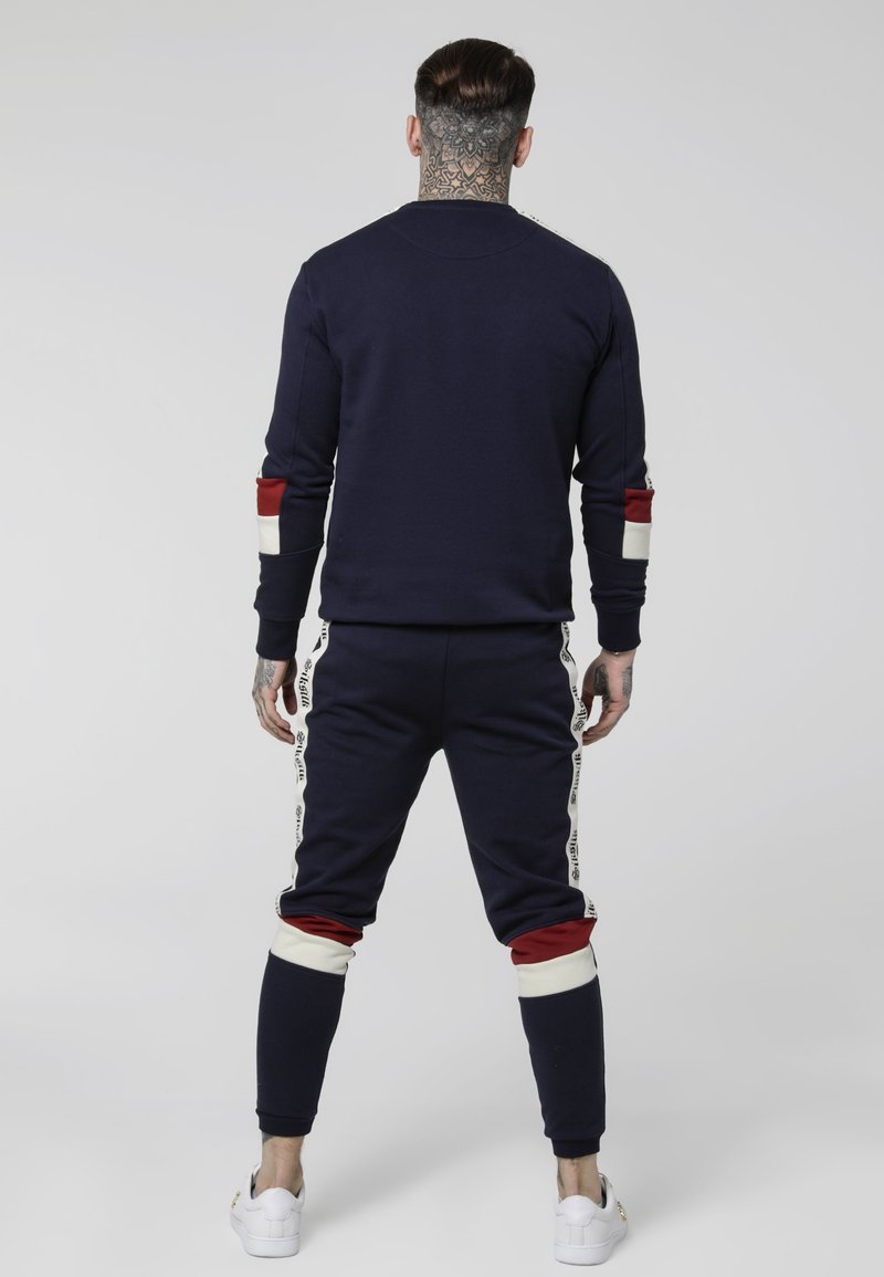 Homme avec des tatouages au cou portant un survêtement bleu marine avec des bandes blanches et rouges sur les manches et le pantalon, debout de dos sur un fond uni.