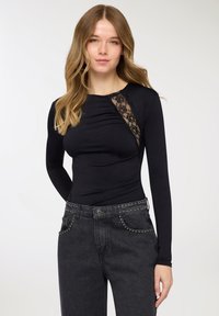 Body a maniche lunghe nero con dettagli in pizzo sulla spalla sinistra, caratterizzato da un design aderente e tessuto strutturato. Abbinato a jeans scuri.