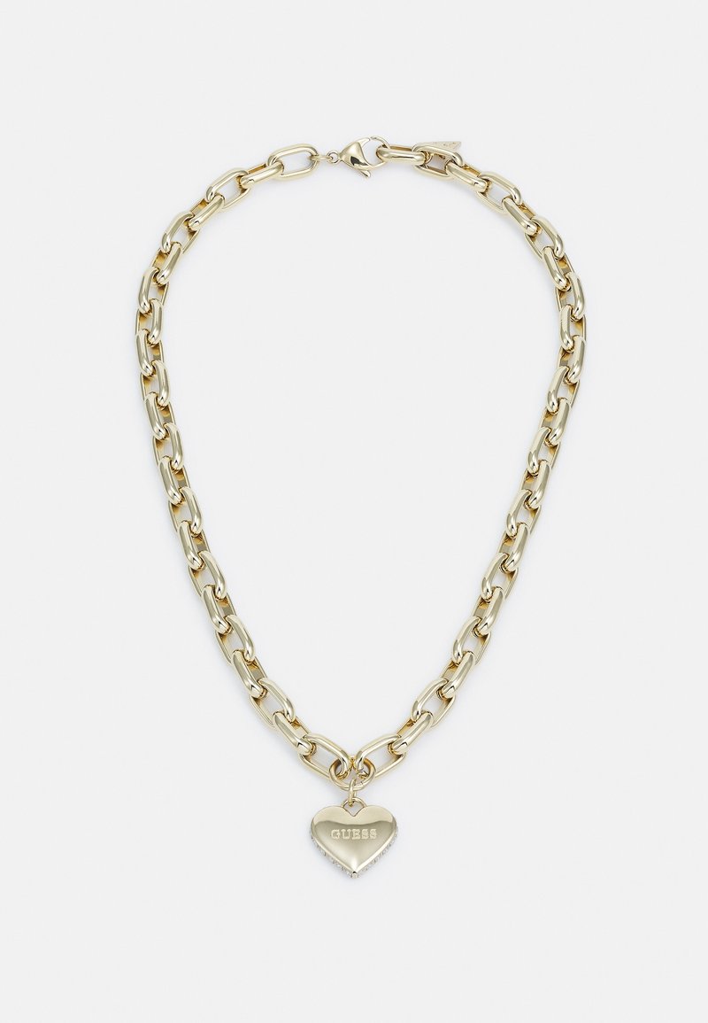 Collier chaîne en or avec un pendentif en forme de cœur gravé du mot "GUESS" sur fond blanc.