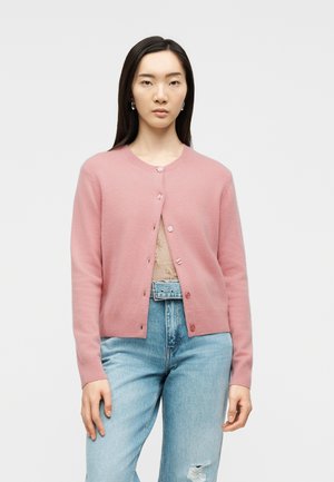 Noor naine kannab roosat nööpidega kardigani beeži topi ja helesiniste kõrge vöökohaga rebitud põlvega teksade kohal, seistes tavalise tausta ees.