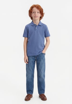 Garçon roux debout de face, portant un polo bleu, un jean bleu et des chaussures marron, une main dans la poche, sur fond blanc.