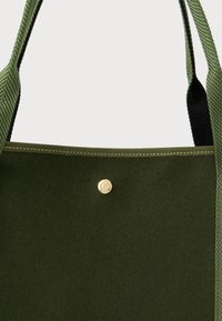 Zelená plátěná taška s zlatým zacvakávacím uzávěrem a kontrastními tmavozelenými popruhy z látky s texturovaným tkaním.