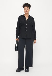 Chemise noire à manches longues avec deux poches avant et fermeture à boutons, associée à un pantalon large noir et des chaussures noires épaisses, tenant un petit sac à main.