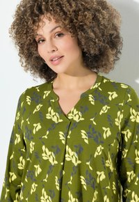 Frau mit lockigem Haar, die eine grüne Bluse mit einem gelb-blauen Blumenmuster trägt und leicht zur Seite schaut, vor einem hellen Hintergrund.