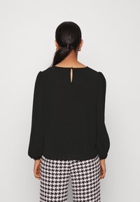 Svart blus med puffärmar som har en nyckelhålsdetalj bak, i kombination med byxor i hundtandsmönster i rosa och svart. Slät textur, avslappnad passform.