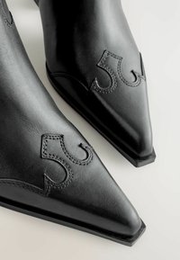 Botas de cuero negras a la altura del tobillo con punta afilada, que presentan un bordado decorativo en forma de onda a los lados y un tacón plano y sólido.