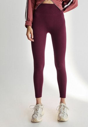 Leggings côtelés bordeaux avec une taille haute, mettant en avant un tissu texturé. Associés à des chaussures de sport de couleur claire pour un contraste.