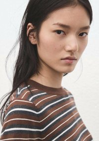 Tânără cu păr lung și închis la culoare, purtând un pulover în dungi maro, gri și alb, privind direct în cameră cu o expresie neutră.