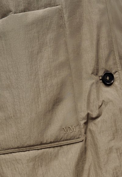 Κοντινή λήψη μπεζ υφασμάτινης τσέπης με κεντημένα αρχικά "MM" και μαύρο κουμπί σε υφασμάτινο ένδυμα με υφή.