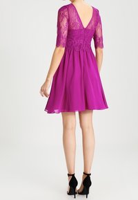 Fialové šifónové šaty s krajkovými rukávy a zadním dílem. Mají hluboký V-zadek, rozšířenou sukni a černé vysoké podpatky.