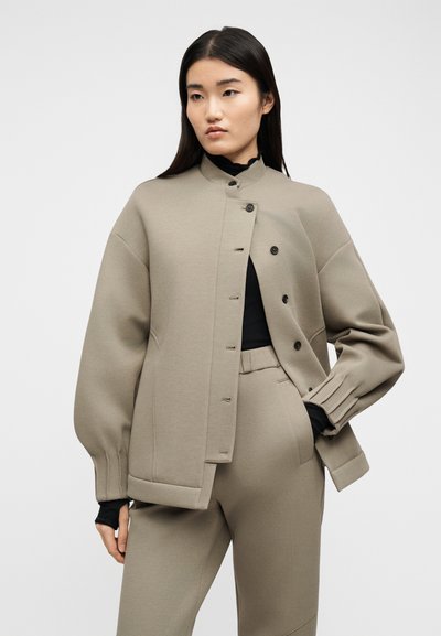 Γυναίκα με φορεμένη μια ταμπά υπερμεγέθη ζακέτα με κουμπιά και ταιριαστό παντελόνι πάνω από μαύρο τοπ, στέκεται μπροστά σε απλό ανοιχτό φόντο.