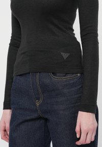 Donna che indossa una maglietta nera aderente a maniche lunghe con un piccolo logo triangolare e jeans blu scuro con cuciture gialle.