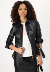 Giacca di pelle nera con design aderente, colletto largo, zip frontale e dettagli sulle spalle; abbinata a una t-shirt bianca con grafica e una gonna di pelle.