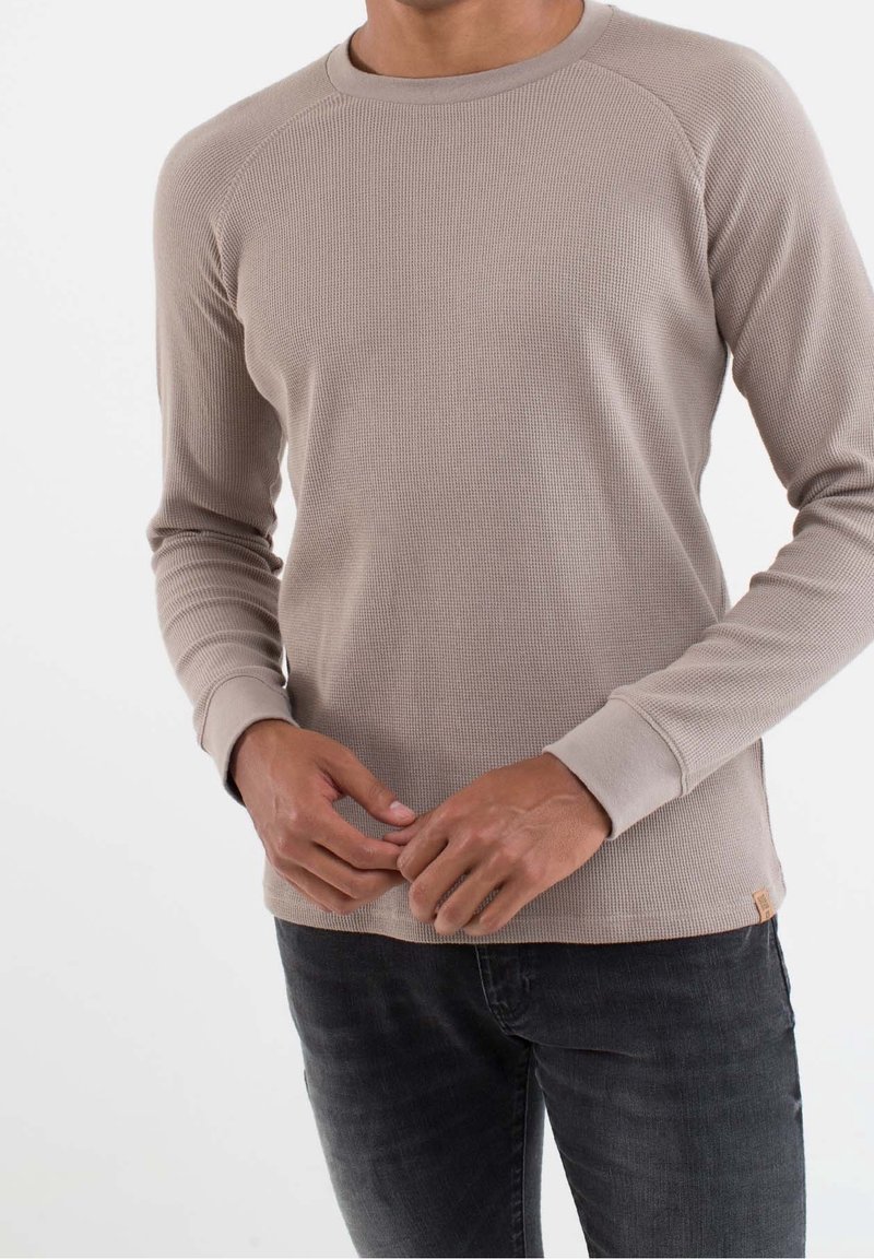 Chemise thermique beige à manches longues avec une texture côtelée, un col rond et des poignets ajustés. Associée à un jean foncé, avec un marquage minimal.