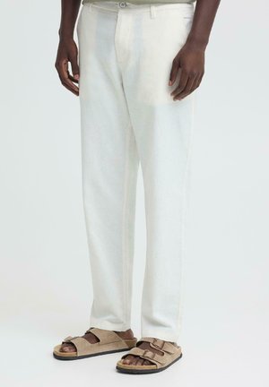 Homme portant un pantalon droit couleur ivoire et des sandales à double bride en suède beige, debout devant un fond uni.