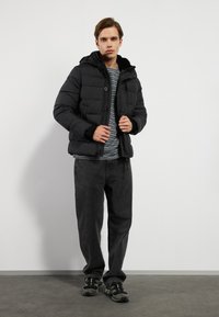 Chaqueta de abrigo negra con capucha desmontable, con textura acolchada, cierre de botones y puños de canalé, llevada sobre una camisa a rayas y pantalones grises.