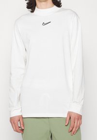 White long-sleeve t-shirt made of cotton, featuring a black Nike logo on the chest, round neckline, and relaxed fit.
