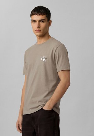 Mladý muž s krátkymi tmavými vlasmi, oblečený v béžovom tričku Calvin Klein a tmavých nohaviciach, stojaci s jednou rukou v vrecku na jednoduchom pozadí.