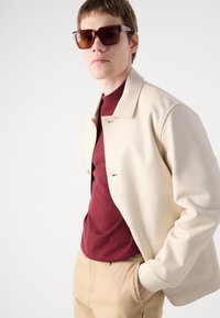 Beige leather jacket with a fold-over collar, over a maroon sweater; model wearing brown square sunglasses, against a light background.
