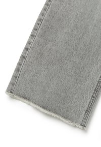 Primer plano de una pierna de pantalón de mezclilla gris con costuras visibles y borde deshilachado sobre fondo blanco.