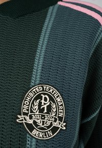 Tissu tricoté vert foncé avec un motif texturé, une bande rose et un patch rond noir et blanc portant l'inscription "Prohibited Textilwaren Berlin 2021-2026."