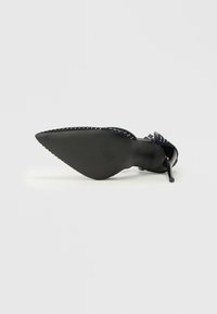 Zapato de tacón alto negro con punta afilada, suela texturizada y tachuelas decorativas a lo largo del borde. El tacón es delgado y de estilo stiletto.