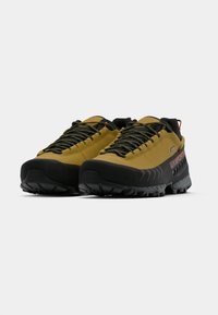 Par de sapatos de caminhada em amarelo mostarda e preto, com solas robustas e atacadores estampados, angulados para mostrar as vistas frontal e lateral.