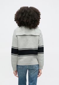 Person mit lockigem Haar trägt einen grauen Pullover mit schwarzen Streifen auf dem Rücken und blaue Jeans mit weißen Textmustern.