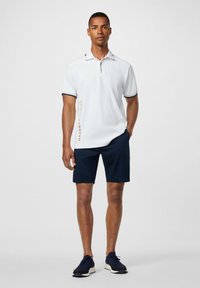 Muž v bielom polokošeli s krátkymi rukávmi a čiernym lemovaním, v námořníckych šortkách, stojaci s jednou rukou v vrecku a neutrálnym výrazom tváre.