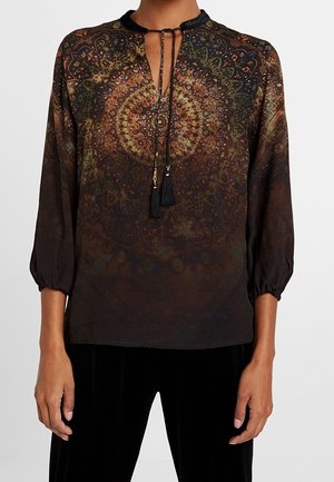 Bluzka z długim rękawem ze ściągaczami, wzór mandali w ziemistych odcieniach, dekolt z kluczem i czarnymi frędzlami, noszona z czarnymi spodniami.