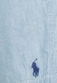 Ljusblå, strukturerad skjorta i tyg med en liten, mörkblå broderad hästlogotyp på vänster sida. Synlig sömdetalj.