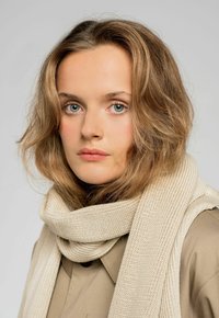 Beżowy dzianinowy szalik nałożony na szyję, z prążkowaną teksturą i miękkim materiałem, dopełniający bluzkę w neutralnym kolorze.