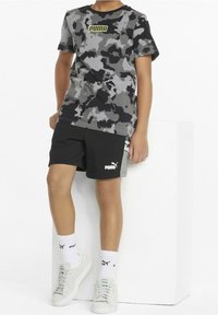 T-shirt grigia a maniche corte con motivo camouflage e logo giallo, abbinata a pantaloni corti neri con logo bianco. Sneakers bianche e calze a coste.