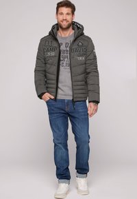 Chaqueta acolchada gris con capucha, con estampados en la parte delantera, combinada con un suéter gris y jeans azules. Zapatillas blancas completan el look.