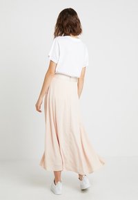 Dlouhá, splývavá broskvová sukně s hladkým texturem, spárovaná s bílým tričkem a bílými teniskami, pohled zezadu.