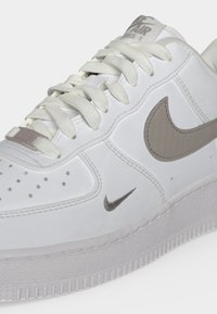 Λευκό παπούτσι Nike Air Force 1 με μπεζ λογότυπα Swoosh, λευκά κορδόνια, οπές στο μπροστινό μέρος και παχιά υφή στη σόλα.