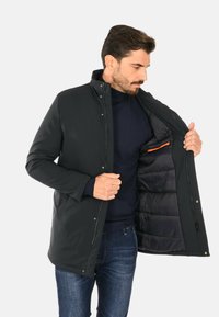 Giacca nera isolante con colletto alto, bottoni a pressione e tasca interna con un accento arancione. Tessuto morbido e liscio con un design elegante.