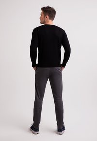 Schwarzer gestrickter Pullover, lange Ärmel, runder Ausschnitt. Graue Slim-Fit-Hose, dezente Textur. Model mit Händen in den Taschen, trägt marineblaue Sneakers.